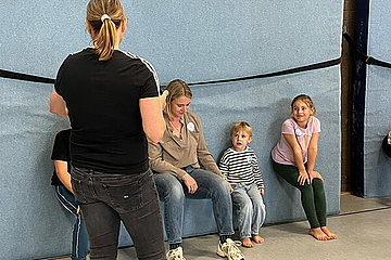 Eine Frau und zwei Mädchen sind halb in der Hocke an eine Wand gelehnt. Übung bei der Kinder-Olympiade. 