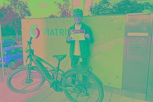 Mann mit Fahrradhelm hält eine MUKOmove-Startnummer in der Hand, vor ihm steht ein Rennrad, im Hintergrund sieht man ein Firmenschild, auf dem Viatris steht. 