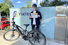Mann mit Fahrradhelm hält eine MUKOmove-Startnummer in der Hand, vor ihm steht ein Rennrad, im Hintergrund sieht man ein Firmenschild, auf dem Viatris steht. 