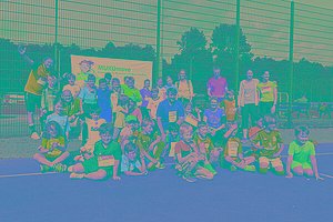 Eine größere Gruppe von Kindern und Erwachsenen sitzt und steht auf einem Sportplatz (roter Schotteruntergrund), viele haben eine MUKOmove-Startnummer angepinnt. 