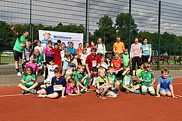 Eine größere Gruppe von Kindern und Erwachsenen sitzt und steht auf einem Sportplatz (roter Schotteruntergrund), viele haben eine MUKOmove-Startnummer angepinnt. 