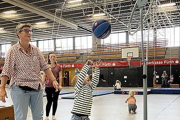 Kleiner Junge versucht, einen Basketball in den Korb zu werfen. Eine Frau steht dabei. 