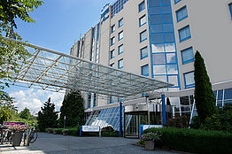 Außenansicht eines mehrstöckigen Gebäudes, wahrscheinlich ein Hotel, mit einem großen Glas-Vordach und einem runden Eingangsbereich