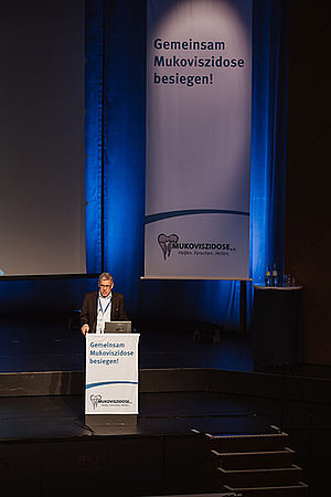 In klein sieht man einen Mann am Podium stehen, im Hintergrund sieht man ein großes Banner des Mukoviszidose e.V.: Stephan Kruip, Vorstandsvorsitzender des Mukoviszidose e.V., eröffnet die Deutsche Mukoviszidose Tagung 2023.