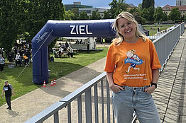 Frau mit orangenem MUKOmove-T-Shirt, in Hintergrund sieht man den Zieleinlauf für eine Laufveranstaltung