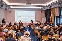Seminarraum mit Zuschauern, vorne sitzen zwei Menschen und simulieren ein Patientengespräch: Dipl. Psych. Cäcilia Siemens und Dr. med. Sandra Barth hielten Impulsvorträge im Rahmen des Workshops des Arbeitskreises Psychosoziales zum Thema: Wie sage ich es? Mit SPIKES? – CF-Behandler als Glücksbringer und Hiobsbotschafter 