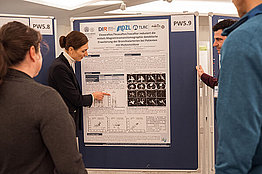 Posterpräsentation auf der DMT 2023, eine Person zeigt auf ein Poster, mehrere Menschen schauen zu. 