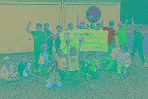 Vor zwei Garagen haben sich mehrere Kinder unterschiedlichen Alters versammelt und posieren mit einem MUKOmove-Banner. 