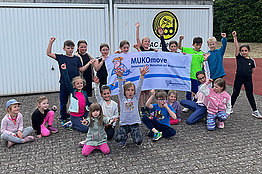 Vor zwei Garagen haben sich mehrere Kinder unterschiedlichen Alters versammelt und posieren mit einem MUKOmove-Banner. 