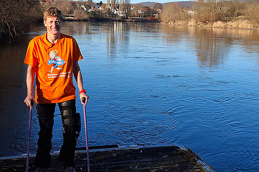 Tina Deeken, Botschafterin des MUKOmove Frau mit Krücken steht vor einem See. Sie trägt ein MUKOmove-T-Shirt.
