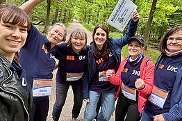 Eine Gruppe von Frauen in Sportkleidung, sie tragen MUKOmove-Startnummern und halten ein MUKOmove Schild in die Luft. Im Hintergrund sieht man Bäume. 