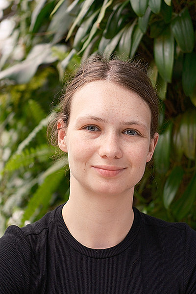 Porträt von Svea Andresen: junge Frau, im Hintergrund sind Bäume und Blätter zu sehen, die Haare sind zu einem Pferdeschwanz gebunden. 