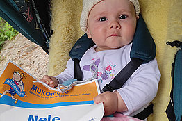 Ein Baby (Mädchen) sitzt im Kinderwagen und hält eine MUKOmove-Startnummer in der Hand, auf der man "Nele" lesen kann. 