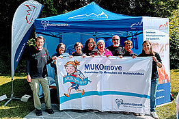 Acht Personen (Männer und Frauen) stehen von einem blauen Pavillon und halten eine MUKOmove-Banner in der Hand. 