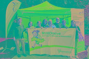 Acht Personen (Männer und Frauen) stehen von einem blauen Pavillon und halten eine MUKOmove-Banner in der Hand. 