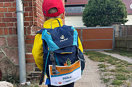 Ein Junge von hinten, er trägt eine gelbe Regenjacke und einen blauen Ranzen. Am Ranzen hängt eine MUKOmove-Startnummer, auf der der Name Mika zu lesen ist. 