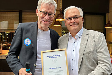 Zwei ältere Männer halten eine Urkunde in die Kamera. 