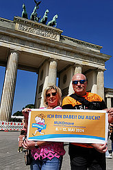 Bei Sonnenschein posieren ein Mann und eine Frau vorm Brandenburger Tor in Berlin mit einem großen Schild, auf dem steht: "Ich bin dabei. Du auch? MUKOmove 2024"