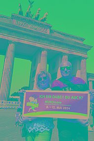 Bei Sonnenschein posieren ein Mann und eine Frau vorm Brandenburger Tor in Berlin mit einem großen Schild, auf dem steht: "Ich bin dabei. Du auch? MUKOmove 2024"
