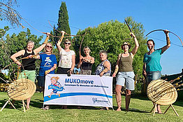 Sieben Menschen halten Bögen in der Hand und stehen draußen bei blauem Himmel neben Zielscheiben. Außerdem halten sie ein MUKOmove-Banner in die Kamera. 