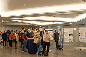 Weiterer Blick in die Posterausstellung, man sieht Gruppen von Menschen, die sich gemeinsam aufgestellte Poster ansehen. 