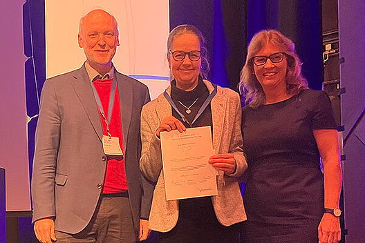 Posterpreis an Dr. Antje Munder Drei Menschen posieren auf einer Bühne, die Frau in der Mitte hält eine Urkunde in der Hand.