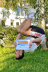 Mann schlägt einen Salto im Garten, er ist über Kopf auf dem Foto zu sehen und hält die MUKOmove-Startnummer richtig herum in die Kamera. 