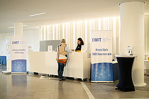 Blick zum Tagungsbüro während der DMT 2023, eine Person steht hinter einem Tresen, eine davor, darum stehen Roll-Ups mit der Aufschrift DMT - Deutsche Mukoviszidose Tagung