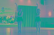 Zwei Tänzerinnen mit schwarzen Kleidern beim Tanzen in einer Tanzhalle, im Hintergrund ist ein MUKOmove-Banner zu sehen. 