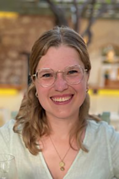 Junge blonde Frau lacht in die Kamera, sie trägt eine Brille, ihre Haare sind lang. Sie trägt ein helles Shirt. 