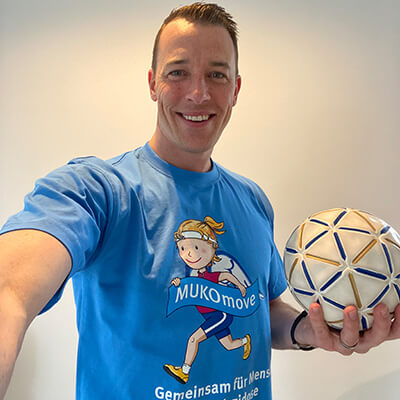 Mann mit blauem MUKOmove-Shirt hat einen Handball in der Hand. Das Foto ist ein Selfie. 