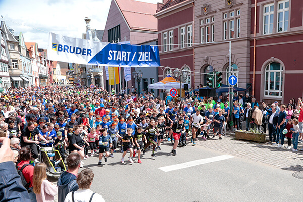 Start einer Laufveranstaltung in einem Stadtzentrum: Man sieht viele Läufer an der Startlinie, darunter auch viele Kinder