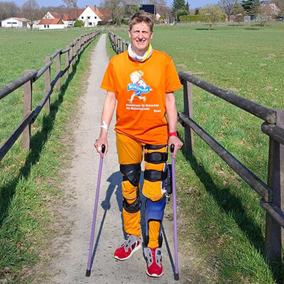 Eine Frau mit Orthesen am Bein und Krücken steht auf einem Feldweg, sie trägt ein orangenes MUKOmove-T-Shirt. 