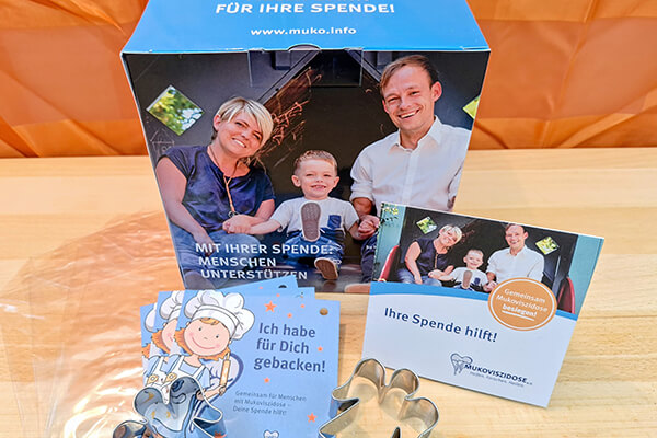 Das Starterpaket für die Schutzengel-Bäckerei Spendenbox aus Pappe, Flyer vom Mukoviszidose e.V., Keksausstechform in Form eines Schutzengels auf einem Tisch mit orangener Decke