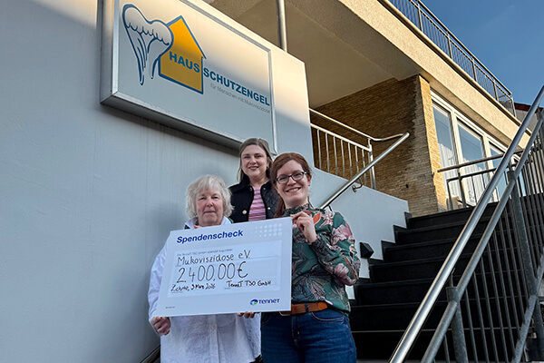 Drei Frauen, eine älter, zwei jünger, halten einen Spendenscheck über 2.400 Euro in der Hand und posieren auf der Treffe eines Hauses, im Hintergrund sieht man ein Logo (Haus mit Schutzengelflügel dazu die Worte "Haus Schutzengel").