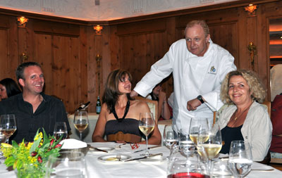 Drei Menschen sitzen an einem festlich gedeckten Tisch, ein Mann in Kochuniform steht.