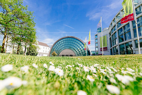 Außenansicht der Stadthalle Bielefeld, man sieht eine Wiese, dahinter die Stadthalle, ein halbrunder Glasbau. 