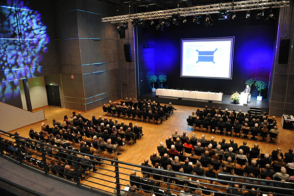 Von oben fotografierter Veranstaltungssaal, man sieht Stuhlreihen und eine Bühne. 
